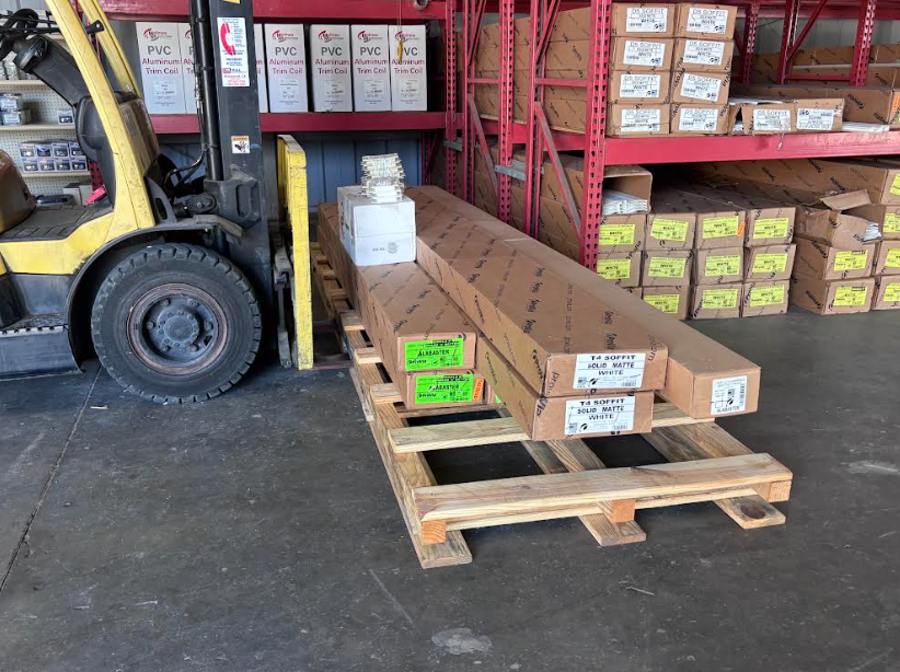Forklift in a warehouse moving long wooden beams on a pallet, with stacked boxes and shelves behind.