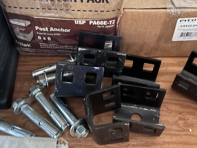 Assorted metal post anchors and bolts scattered on a wooden surface in front of cardboard boxes.