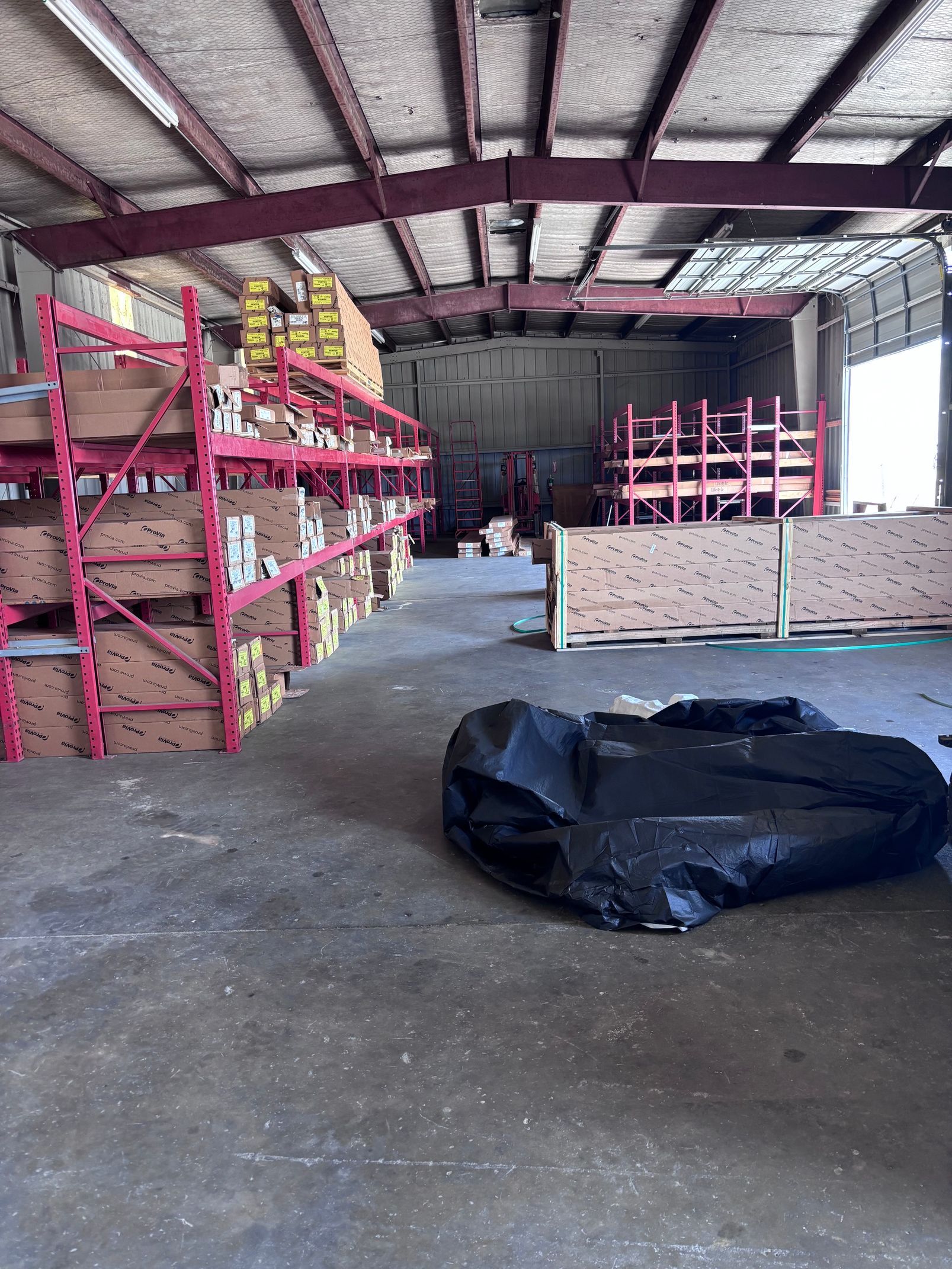 Warehouse interior with pink shelving, stacked supplies, and a black tarp-covered bundle on the floor