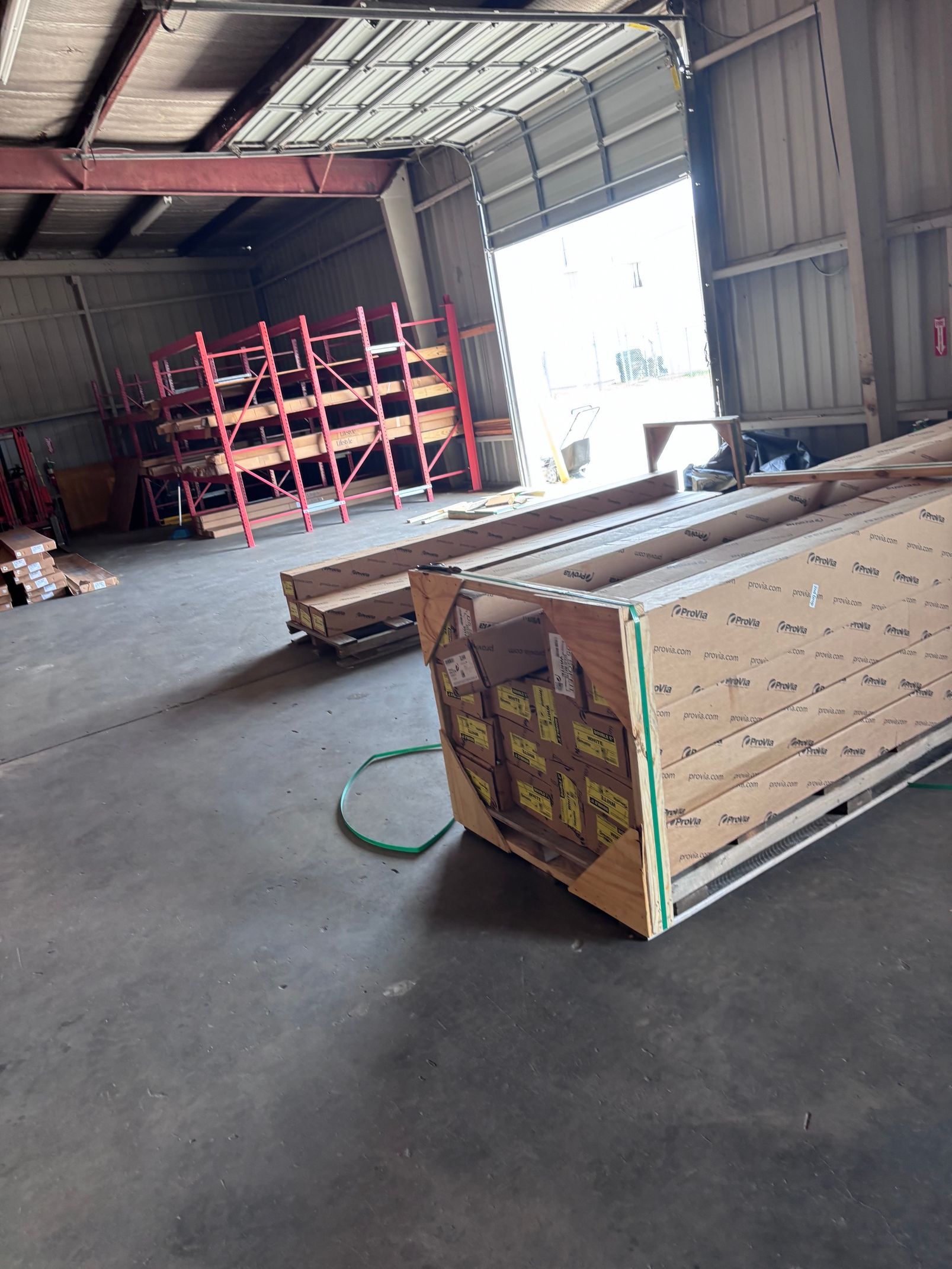 Large wooden crate on a warehouse floor beside red shelving and a green hose, with a bright loading bay in back