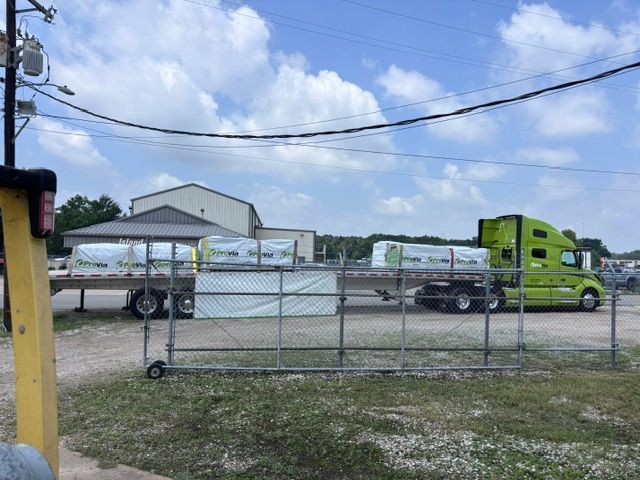 Green semi truck and white tanker trailer behind a chain-link fence at an industrial site under a blue sky.
