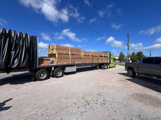 Flatbed trailer loaded with stacked lumber in a gravel lot under a blue sky.