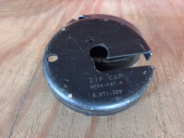Black ZIP CAP with a keyhole and mounting hole, lying on a wooden surface.
