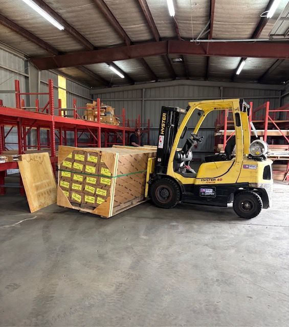 Yellow forklift in a warehouse, moving large wooden crates beside red shelving.