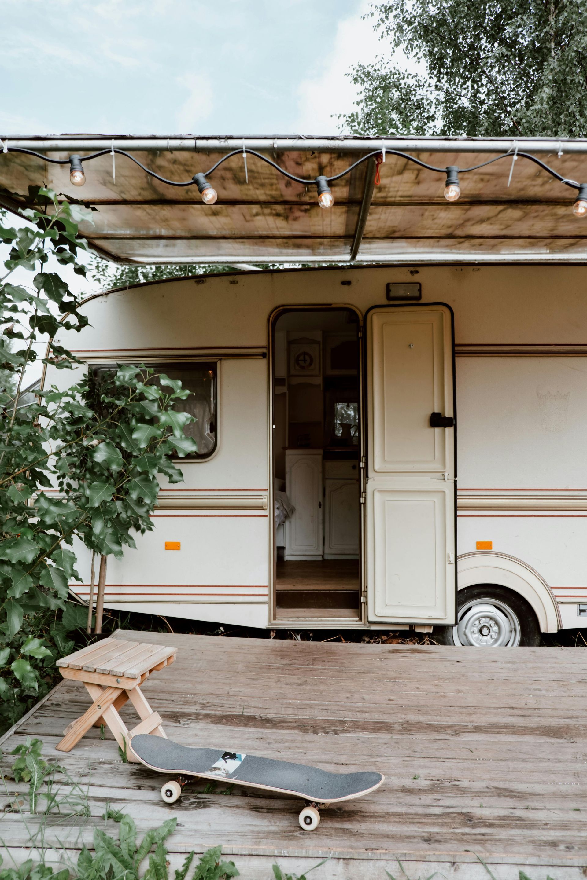 A vintage camper trailer with an open door and string lights sits on a wooden deck with a small stool and a skateboard.