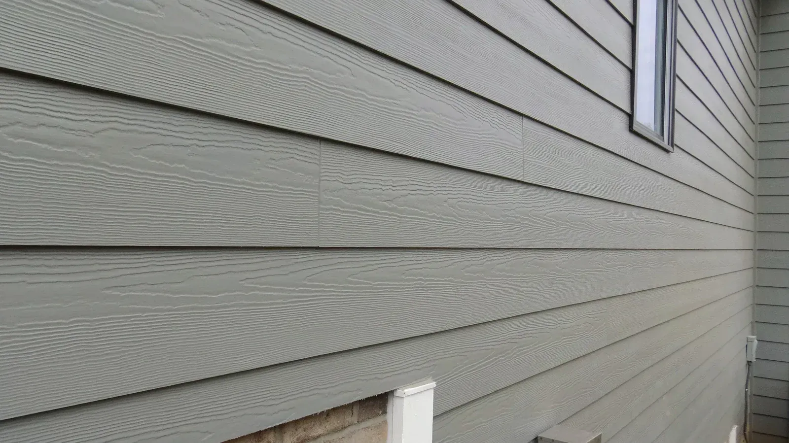 Grey, wood-grain patterned horizontal lap siding on the side of a building, showing a window and a corner.