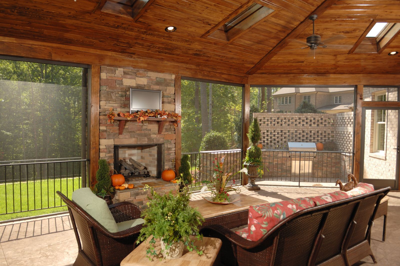 Covered patio with wood ceiling, dining table, wicker chairs, fireplace, and large windows overlooking greenery