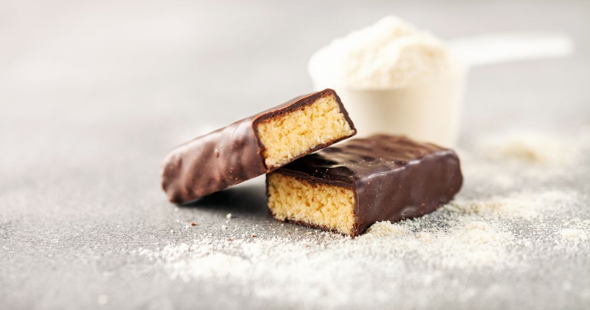 A chocolate bar with a bite taken out of it is sitting on a table next to a scoop of protein powder.