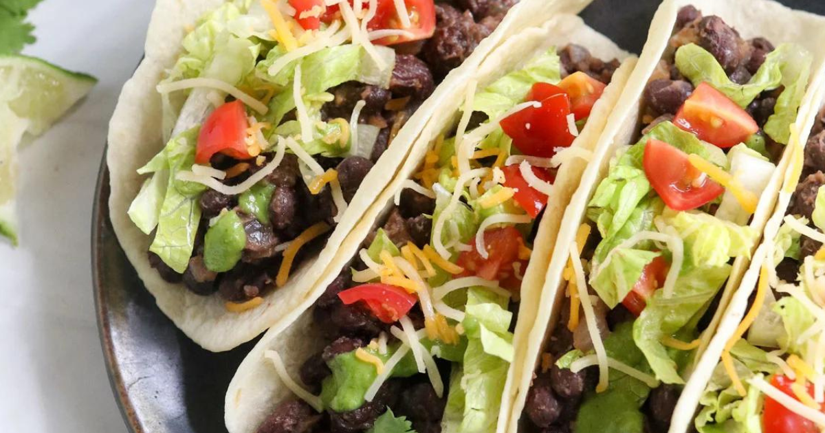 A close up of three tacos with black beans , lettuce , tomatoes and cheese on a plate.