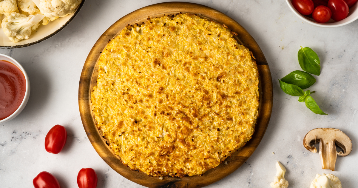 A cauliflower pizza crust is sitting on a wooden cutting board next to tomatoes and mushrooms.