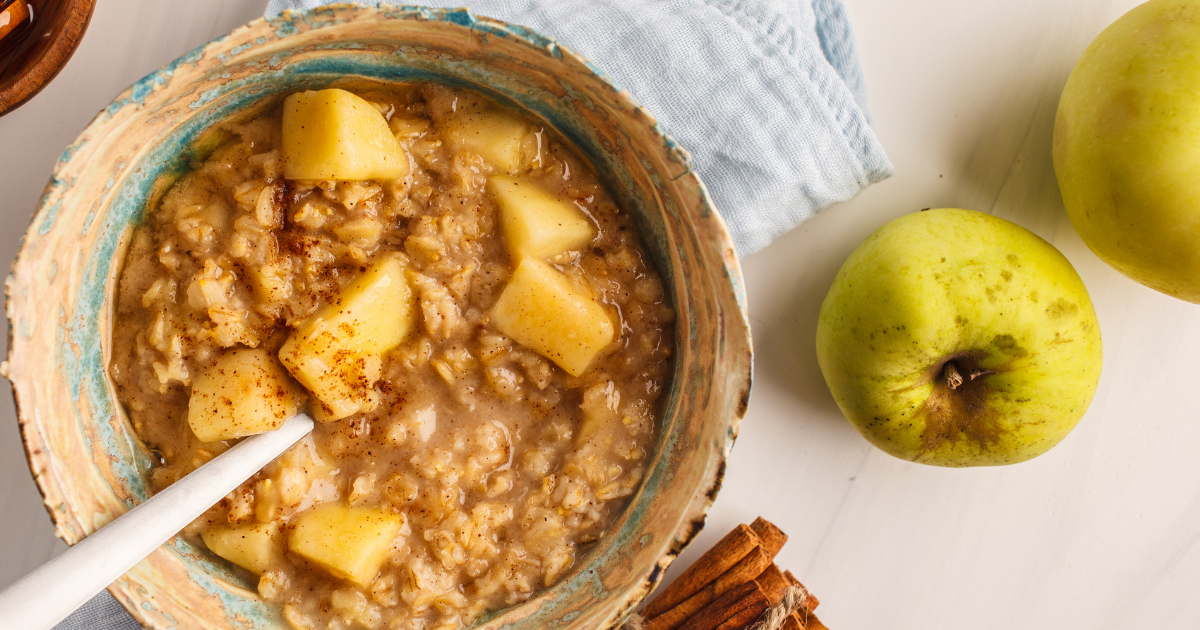 A bowl of oatmeal with apples and cinnamon next to apples and cinnamon sticks.