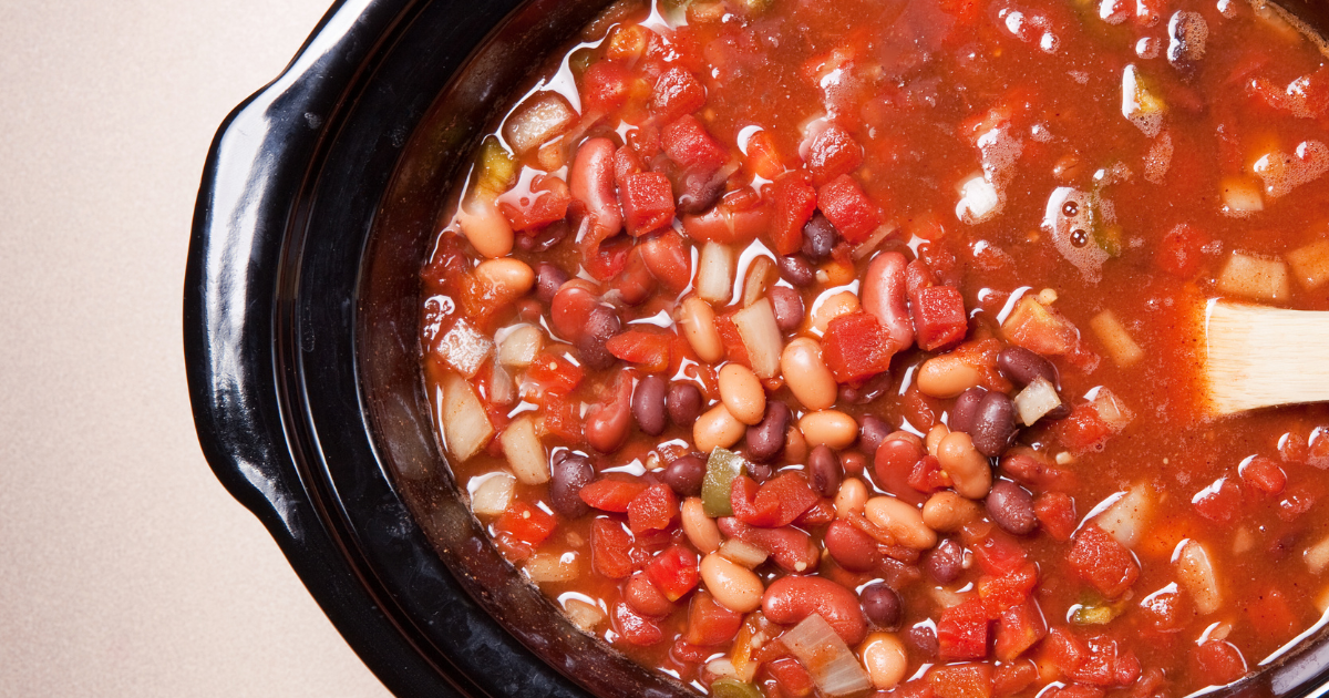 A crock pot filled with beans and tomatoes with a wooden spoon in it.
