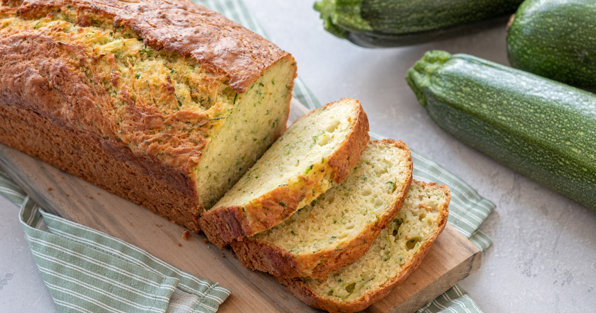 A loaf of zucchini bread is sitting on a wooden cutting board next to zucchini.