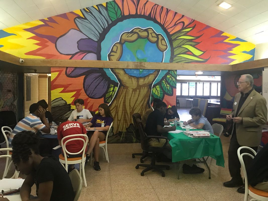 People at tables in a room with a mural of a hand holding the earth. A man in a suit is near the tables.