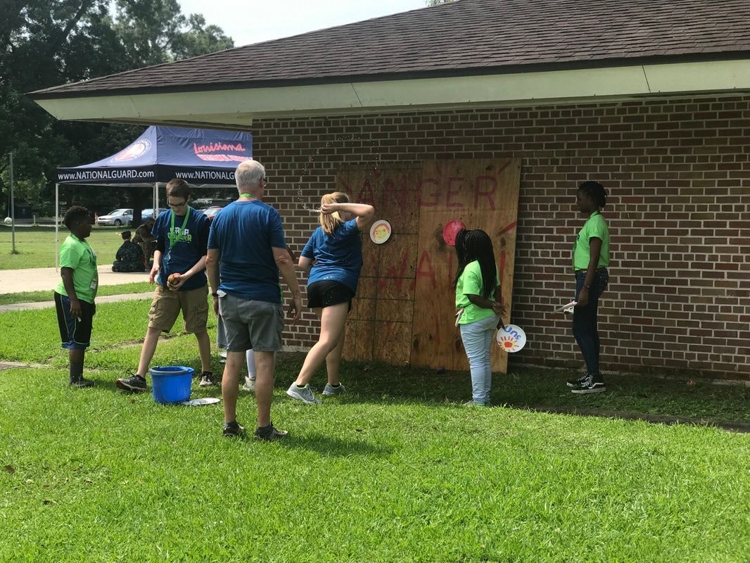 People playing a game tossing objects at a board on a brick building, outdoors on a sunny day.