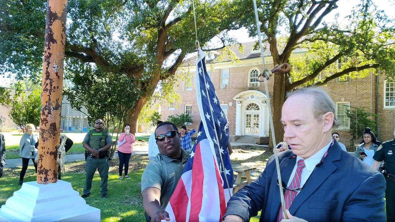 People raising a U.S. flag on a pole outdoors, historic building in background.