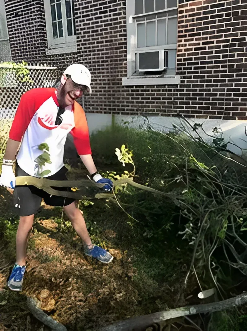 Man in red and white shirt, white cap, holding branches, smiling, working outdoors near a brick building.