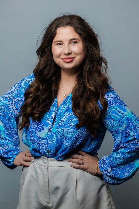Woman in a blue patterned blouse and light gray pants, standing with hands on hips and smiling against a gray backdrop.