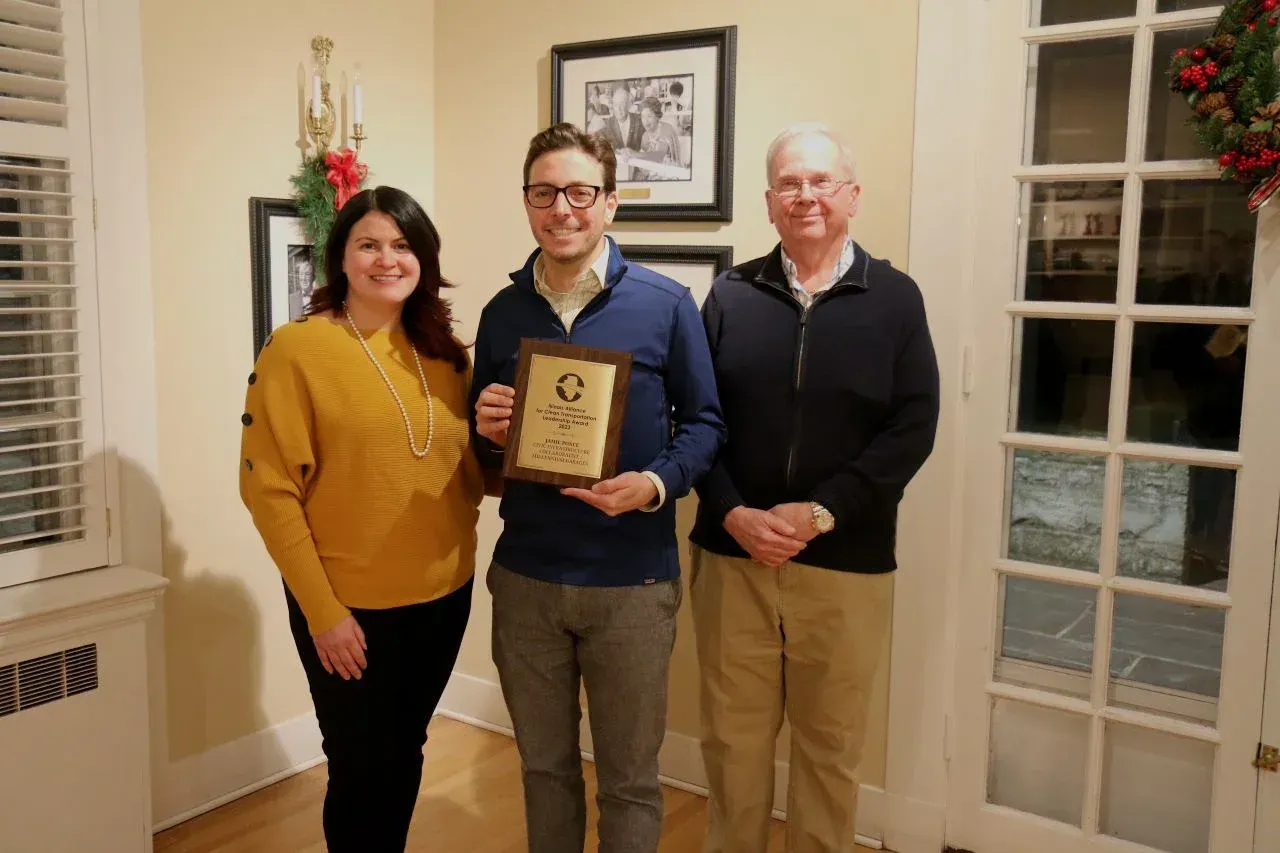 Three people pose for a photo, one holding an award. Woman in yellow, man in blue, and older man in beige stand inside a building.