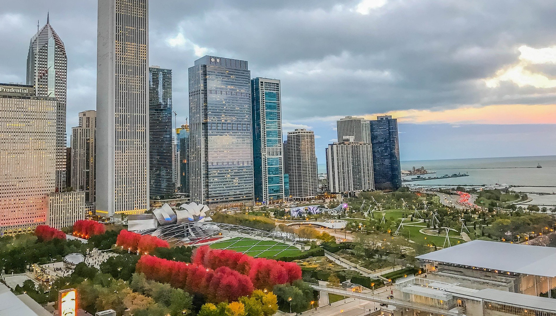 Cityscape with modern skyscrapers, park, and water under a cloudy sky.