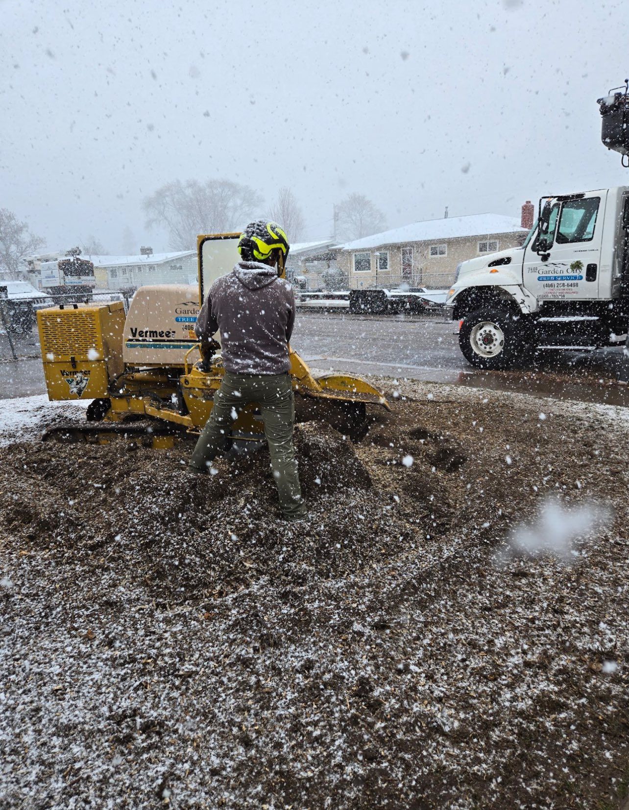 A person operates a stump grinder in the snow, surrounded by wood chips. A truck is parked nearby in a residential area.