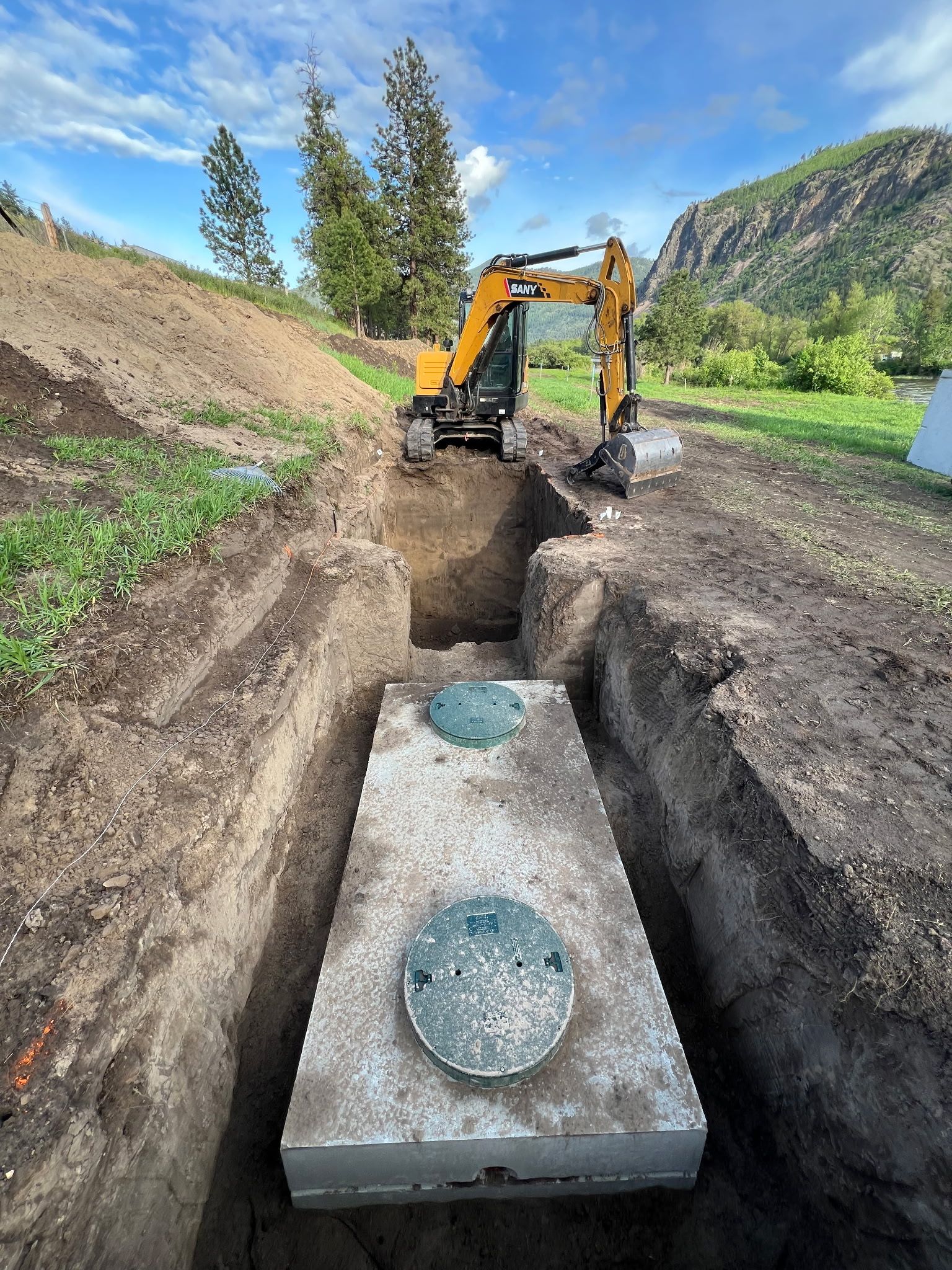 An excavator is working to bury a concrete septic tank in a deep trench. Green lid visible. Mountains and blue sky in the background.