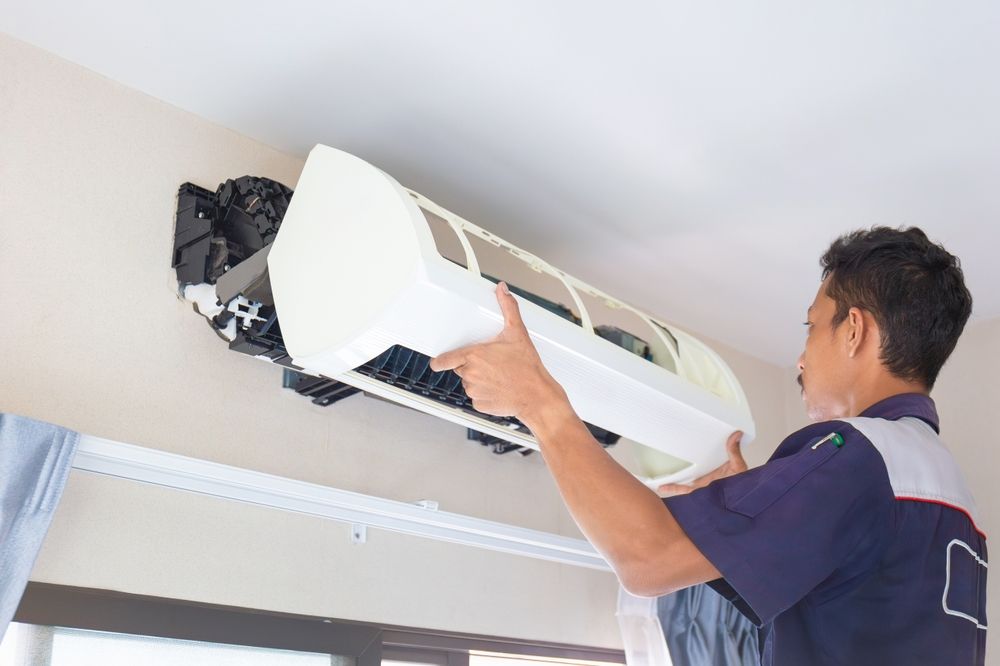 A technician wearing a work uniform installs a white wall-mounted air conditioning unit in an indoor room.