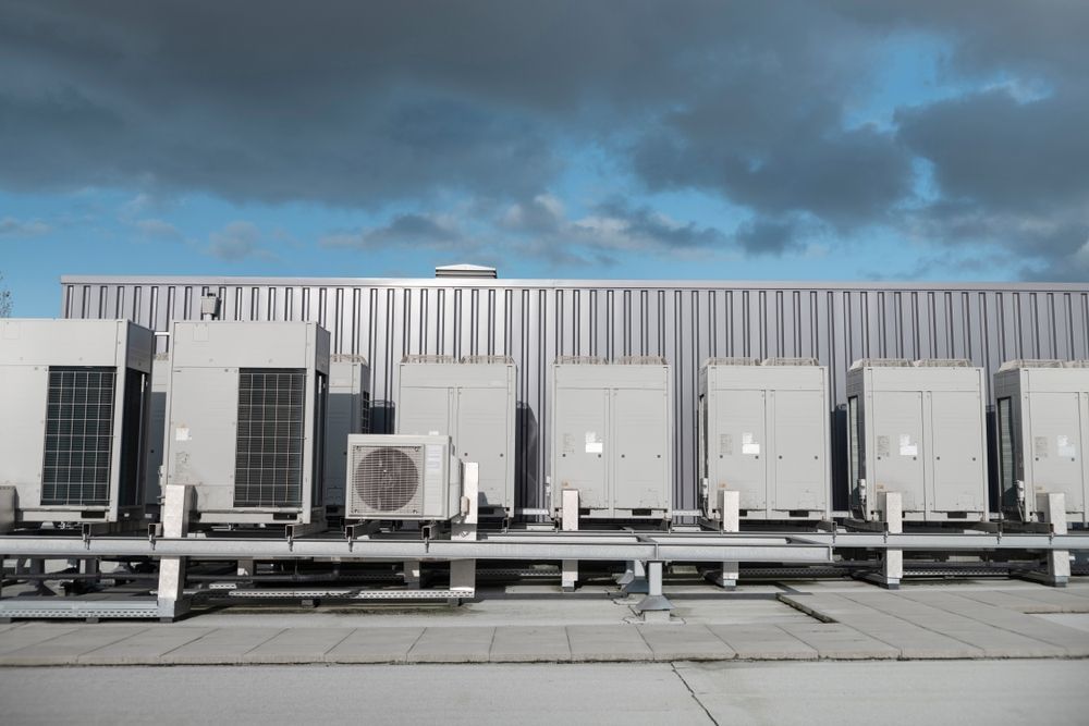 A row of industrial air conditioning units sits on a rooftop against a blue sky with grey clouds.