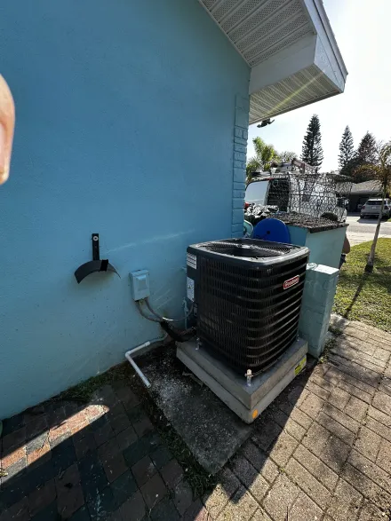 An outdoor HVAC condenser unit sits on a concrete pad against the blue stucco wall of a house, beside a brick walkway.