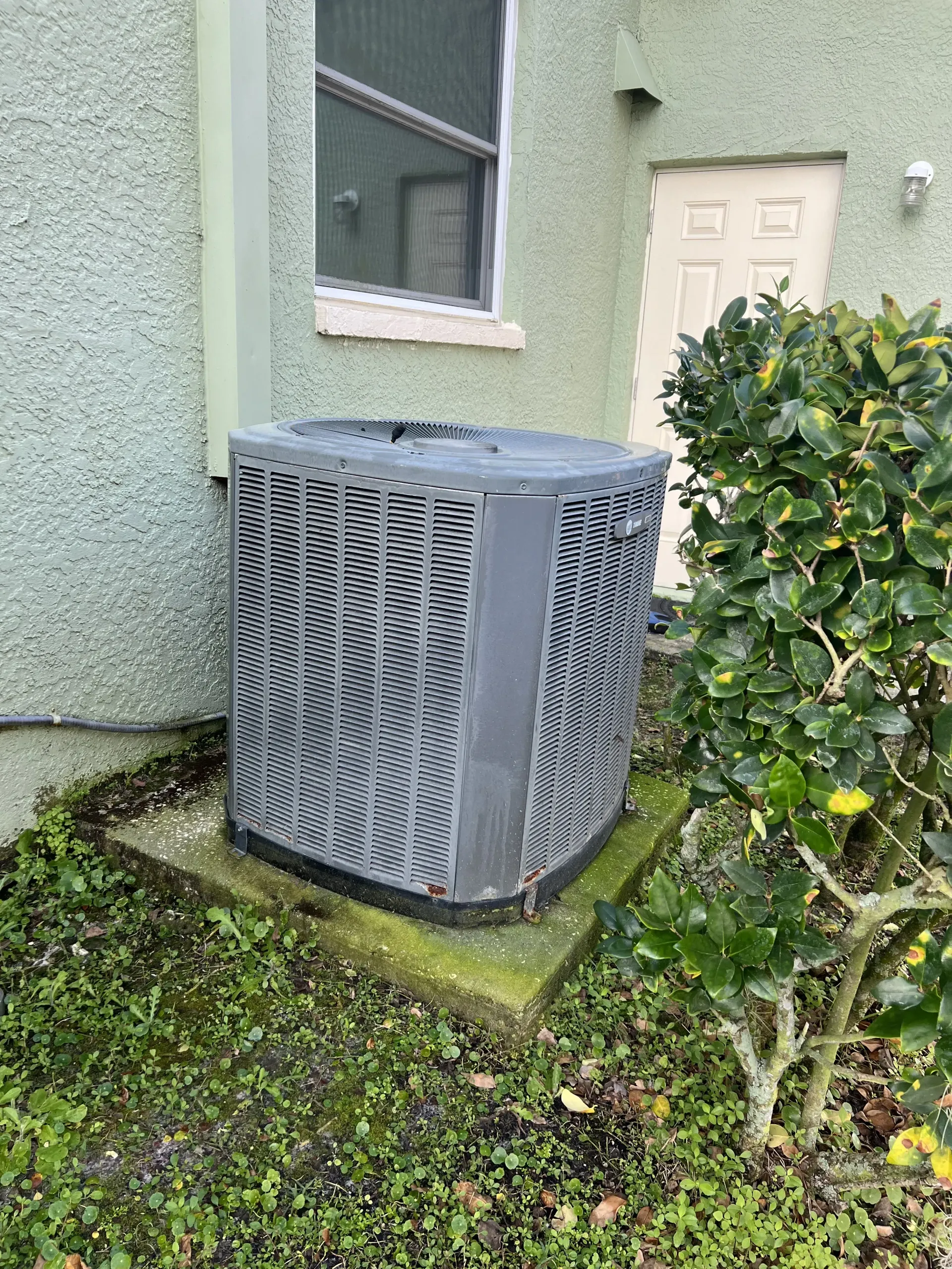 A grey, rectangular residential air conditioning unit sitting on a concrete pad outside a light-colored stucco building.