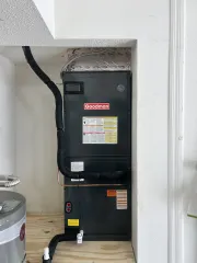 A Goodman HVAC air handler unit standing in a small utility closet on a plywood floor next to a water heater.