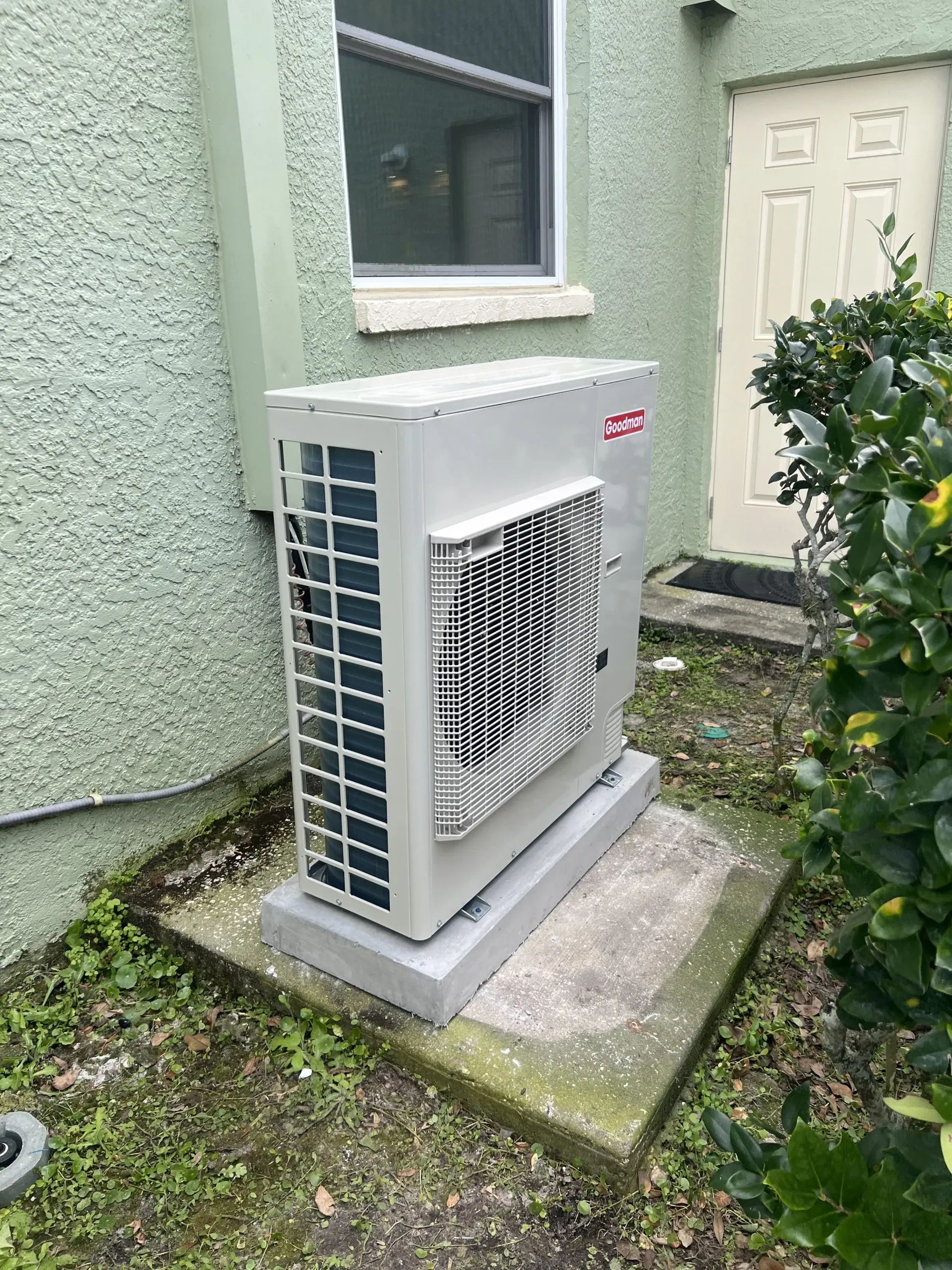 A light-colored outdoor air conditioning condenser unit sitting on a concrete pad against a green stucco house wall.