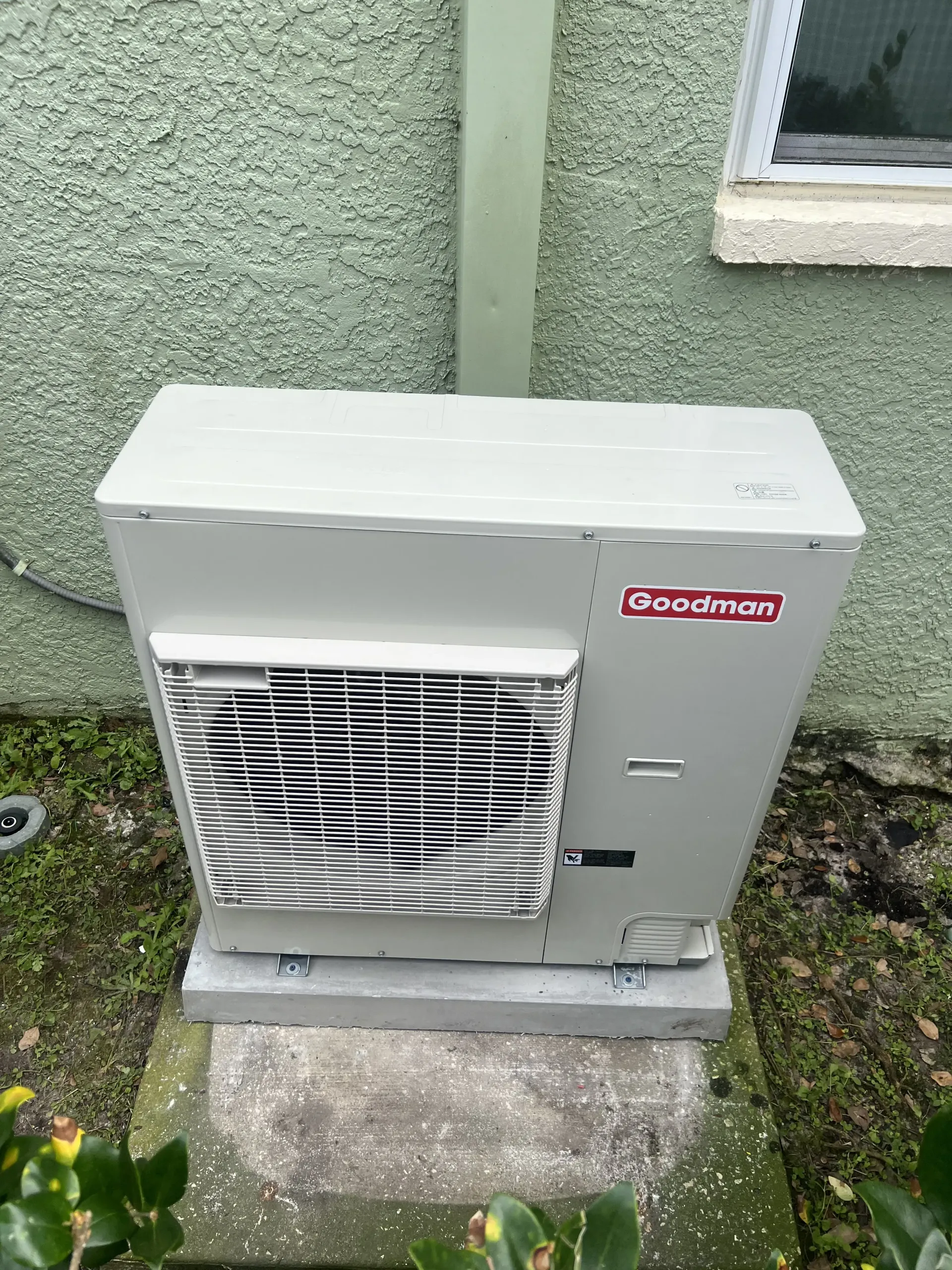 A beige Goodman outdoor air conditioner unit sitting on a concrete pad against a green stucco wall.