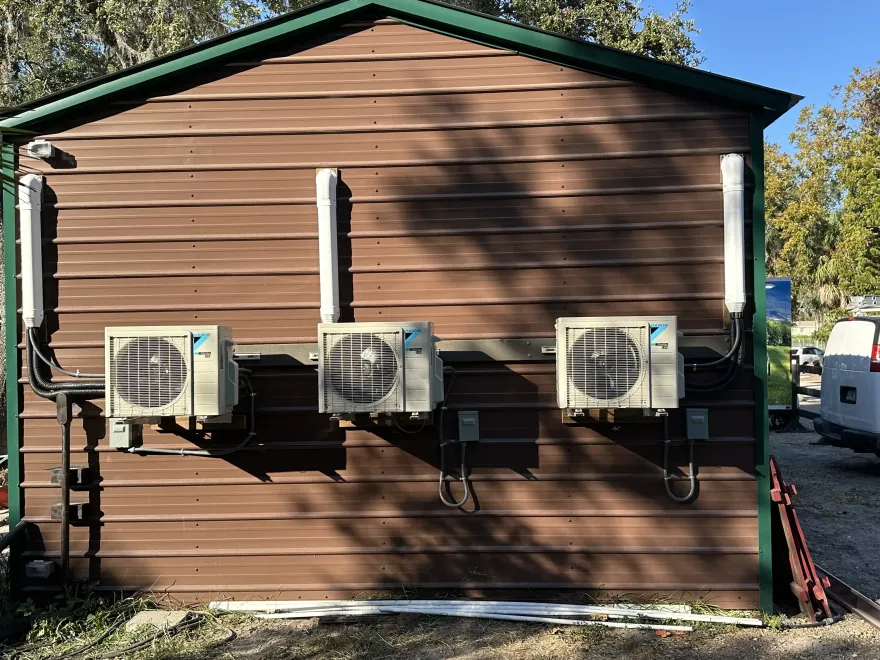 Three HVAC units mounted on the exterior brown wall of a building with visible vertical white pipe covers.