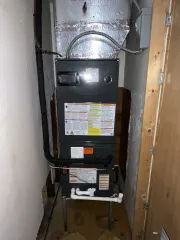 A dark gray HVAC unit standing in a closet alcove, with metal ductwork above and white drainage pipes below.