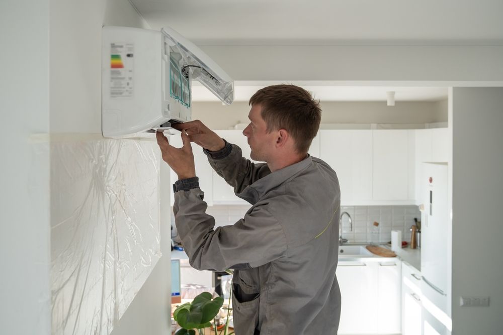 A technician in a grey work uniform repairs a white wall-mounted air conditioner in a bright, modern kitchen.