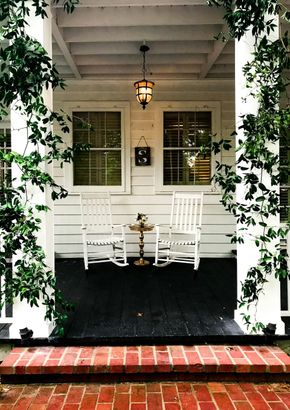 A porch with two white rocking chairs, a small table, and a lantern, framed by climbing ivy and brick steps.