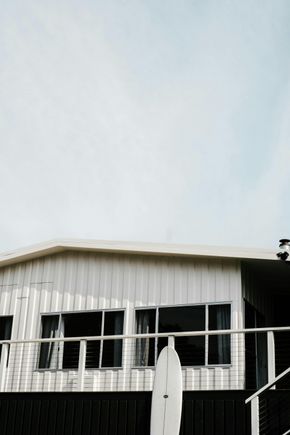 A light-colored, single-story building with vertical siding and large windows, partially obscured by a white surfboard.