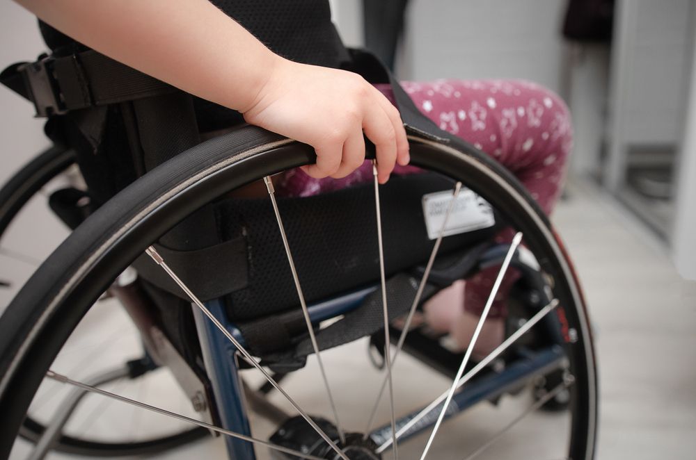 Child's hand on a wheelchair wheel. Person in patterned pants visible. Indoor setting. — Bourbong Street Physiotherapy Centre in Bundaberg West, QLD