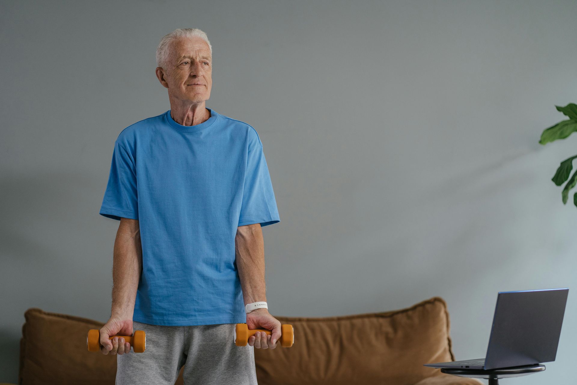 An older person in a blue shirt and gray pants lifts dumbbells at home in front of a laptop. — Bourbong Street Physiotherapy Centre in Bundaberg West, QLD