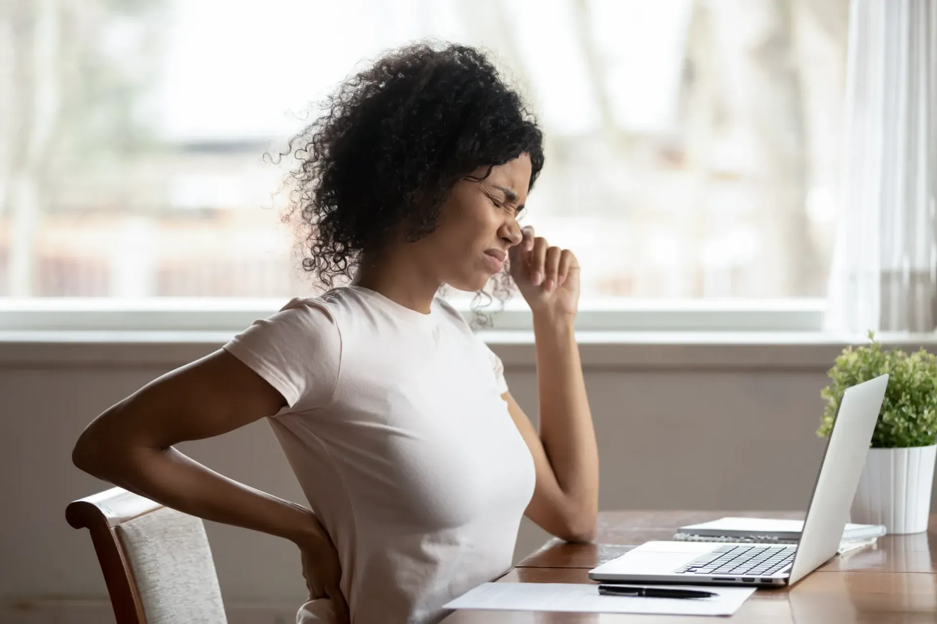 Woman at desk with laptop, grimacing while touching her eye and lower back; indoors. — Bourbong Street Physiotherapy Centre in Bundaberg West, QLD