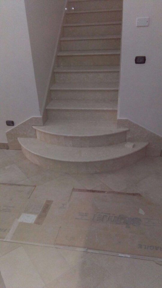 Vista interna di una scala in pietra beige con due gradini inferiori curvi che conducono a una rampa di scale rettilinea.