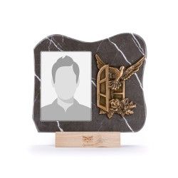 Una targa commemorativa in marmo con uno spazio vuoto per un ritratto, accanto a una colomba in bronzo e a un bassorilievo su una finestra, su una base di legno.
