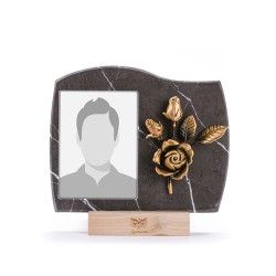 Una targa commemorativa in marmo scuro con una fessura verticale per la foto e una scultura a forma di rosa in ottone, poggiata su una base di legno.