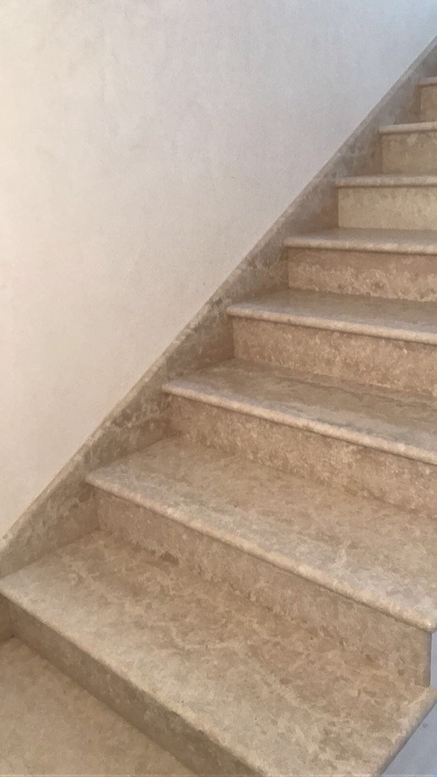 Una scala con gradini in pietra beige che conducono verso l'alto, addossata a una parete semplice e di colore chiaro.