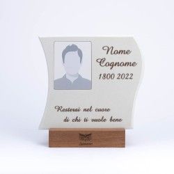 Una targa commemorativa con ritratto, nome e date provvisori, posta su un supporto di legno su sfondo bianco.