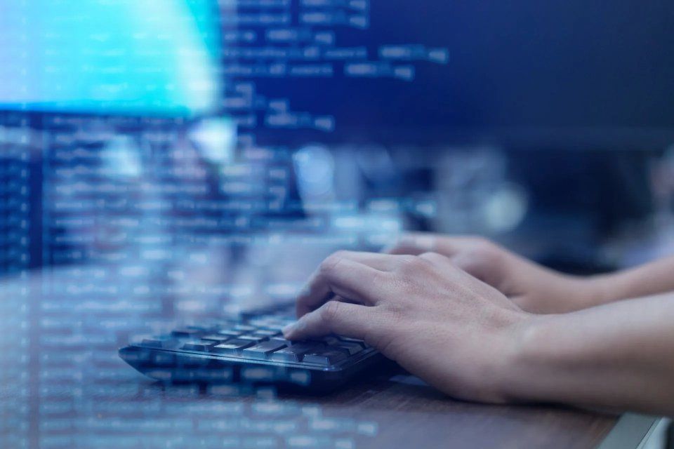 Mani che digitano su una tastiera; codice visualizzato sullo schermo di un computer sullo sfondo.