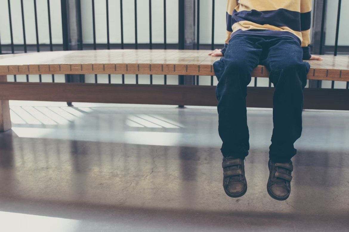 Person in jeans sits on a bench in a bright room, legs dangling over a polished floor.
