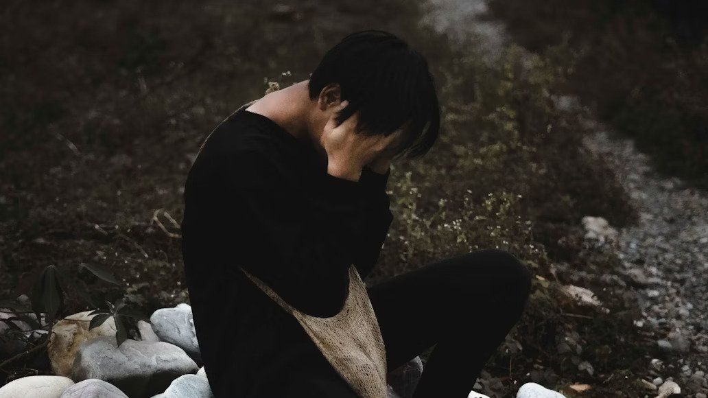 Person in black clothing sits on rocks, face hidden in their hand, in a dark outdoor setting.
