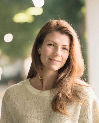 Smiling woman in a cream sweater with long hair, standing outdoors in soft sunlight.